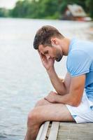Feeling lonely and depressed. Side view of depressed young man touching head with hand and looking down while sitting at the riverbank photo