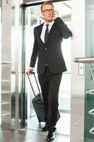 Executive on the go. Full length of confident young man in formalwear carrying suitcase and talking on the mobile phone while getting out of elevator photo