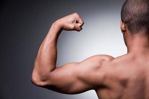 orgulloso de su cuerpo perfecto. vista trasera de un joven africano sin camisa mirando su bíceps mientras se enfrenta a un fondo gris foto