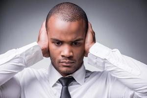 Hear no evil. Young African man in shirt and tie covering ears with hand while standing against grey background photo