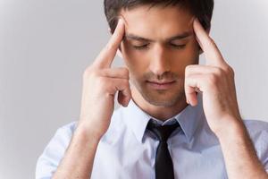 He got stressful job. Young man in shirt and tie touching head with fingers and keeping eyes closed while standing against grey background photo