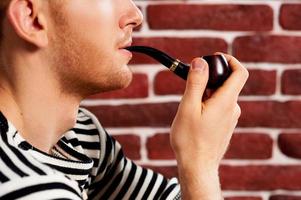 Smoking a pipe. Close-up of young man in striped clothing smoking pipe while sitting on the chair photo