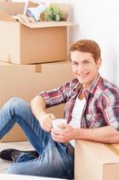 Just moved to the new apartment. Top view of cheerful young man sitting on the floor of his new apartment and drinking coffee while cardboard boxes laying in the backgroundJust moved to the new apartment. Top view of cheerful young man sitting on the floo photo