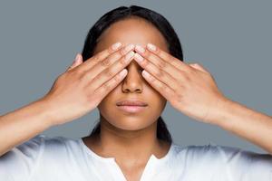 no veo nada. bella joven africana cubriendo los ojos con las manos mientras se enfrenta a un fondo gris foto