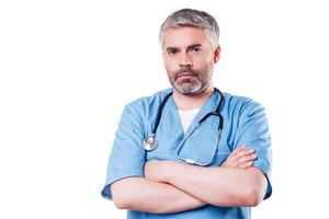 Confident surgeon. Mature grey hair doctor looking at camera and keeping arms crossed while standing isolated on white photo