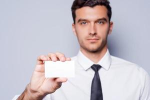 Copy space on his business card. Confident young man in shirt and tie showing his business card while standing against grey background photo