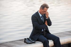 Feeling lonely and depressed. Depressed mature businessman touching his face with hand while sitting barefoot at the quayside photo