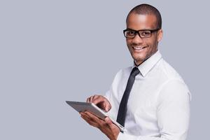 empresario con tableta digital. un joven africano confiado con camisa y corbata trabajando en una tableta digital y sonriendo mientras se enfrenta a un fondo gris foto