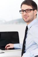 Confident businessman. Rear view of cheerful young man in shirt and tie looking over shoulder while sitting at his working place photo