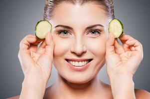 Natural spa treatment. Beautiful mature woman holding pieces of cucumber near her eyes and smiling while standing isolated on white background photo