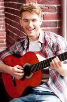 Singing his favorite song. Handsome young man playing guitar and singing while sitting on the window sill photo