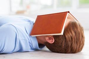 libro aburrido. vista lateral de un joven con un libro en la cara tendido en el suelo de su apartamento foto