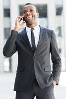 comunicaciones de negocios. feliz joven africano con ropa formal hablando por teléfono móvil y sonriendo mientras está de pie al aire libre foto