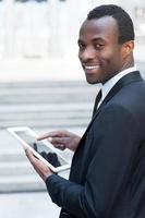 Working on tablet. Cheerful young African man in formalwear working on digital tablet and looking over shoulder while standing outdoors photo