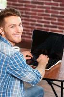 Surfing the net is fun. Top view of handsome young man working on laptop and looking over shoulder while sitting at his working place photo