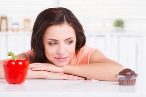 Sweet temptation. Thoughtful young woman choosing what to eat while leaning at the kitchen table with chocolate muffin and fresh pepper laying on it photo