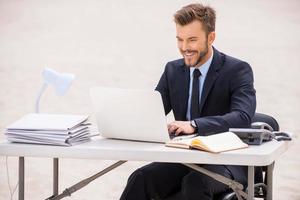 Finding a peaceful place to work. Cheerful young man in formalwear working on laptop while sitting at the table on sand photo