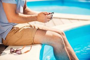 Chatting with friends. Close-up side view of man sitting poolside and typing something on mobile phone photo