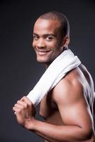 After workout. Young shirtless African man carrying towel on shoulders and looking over shoulder with smile while standing against grey background photo