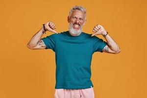 Happy senior man looking at camera and pointing himself while standing against orange background photo