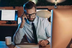 Confident young man working on computer and talking on mobile phone while staying late in the office photo