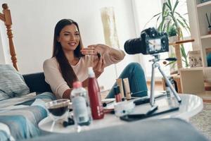 Beautiful young woman pointing at beauty product and smiling while making social media video photo