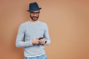 Handsome young man in casual wear using smart phone and looking at camera while standing against brown background photo