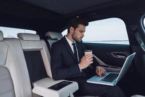 Business on the go. Handsome young man in full suit working using laptop while sitting in the car photo