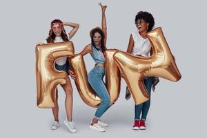 Full length of three attractive young women carrying balloons and smiling while standing against grey background photo