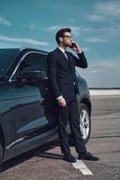 Talking about business. Full length of handsome young businessman talking on the phone while standing near his car outdoors photo