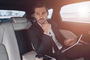 joven perfeccionista. un joven apuesto con traje completo que trabaja con una tableta digital mientras está sentado en el auto foto
