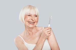 Taking good care of teeth. Beautiful senior woman holding toothbrush and smiling while standing against grey background photo