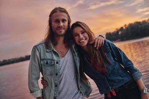 They belong together. Happy young couple in casual wear smiling and looking at camera while standing near the lake photo