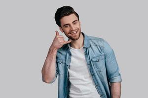 Give me a call Good looking young man in blue jeans shirt gesturing and looking at camera while standing against grey background photo