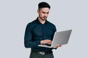 Using laptop.  Handsome young man in shirt working on laptop while standing against grey background photo