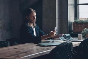 Sending message.  Handsome young man in smart casual wear holding smart phone and looking at it while sitting at his desk in office photo