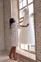 Exited about her wedding. Full length of attractive young woman in silk bathrobe touching her wedding dress while standing near the window photo