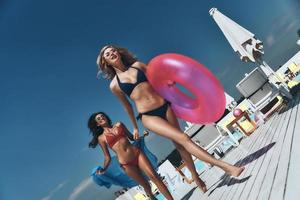 Having great time together. Two beautiful young women in bikini smiling while running near the pool together photo