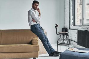 He is looking into your heart. Full length of thoughtful young man in white shirt and suspenders looking at camera and keeping hand on chin while leaning on the sofa photo