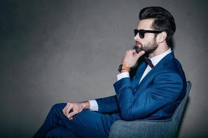 All about style. Side view of young handsome man in suit and bow tie holding hand on chin and looking away while sitting in grey chair against grey background photo