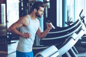 Moving fast. Side view of young man in sportswear running on treadmill at gym photo