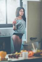 Only the freshest veggies for my salad. Beautiful young African woman standing near the fridge and holding red pepper while standing in kitchen at home photo