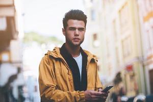 City style. Handsome young man holding smart phone and looking away while standing on the city street photo