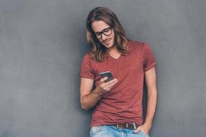 Typing quick message to friend. Handsome young man in casual wear holding smart phone and looking at it while standing against grey background photo
