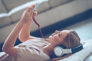 la música le alegra el día. vista lateral de una hermosa joven escuchando música mientras está acostada en la alfombra en casa foto