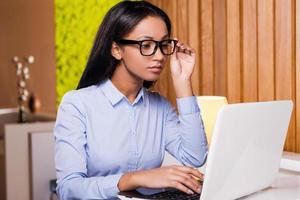 Confident businesswoman. Attractive young African woman working on laptop and adjusting her eyeglasses while sitting at her working place photo