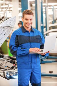 Full of confidence and expertise. Confident young man working on laptop and smiling while standing in workshop with car in the background photo