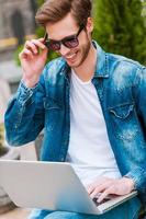 Enjoying free Wi-Fi. Cheerful young man working on laptop and adjusting his eyewear while sitting outdoors photo