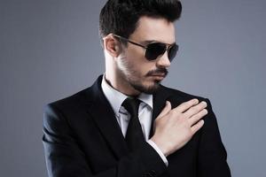 Used to perfection and success. Handsome young man in formalwear and sunglasses adjusting his jacket while standing against grey background photo