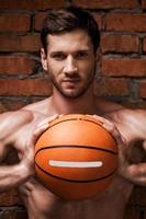 He is willing to this game. Handsome young muscular man holding basketball ball while standing against brick wall photo